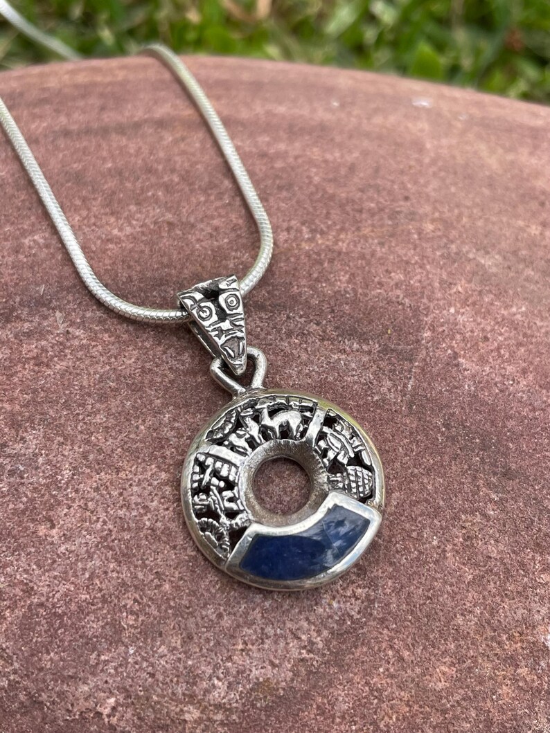 May include: A silver pendant necklace featuring a circular pendant with intricate carvings and a dark blue stone inlay. The pendant hangs from a delicate silver chain, showcasing a detailed design with a central hole.