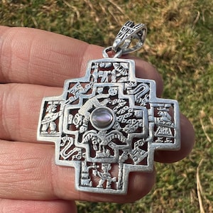 May include: A silver-toned metal pendant in the shape of a stepped cross, featuring intricate cut-out relief carvings of figures, animals, and geometric patterns, with a small, round, iridescent cabochon stone set in the center of the design.