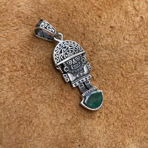 Puede incluir: Colgante de plata con un diseño intrincado de una figura estilizada con un tocado y una piedra preciosa verde. El colgante cuelga de una fianza decorativa. El fondo es una superficie marrón.