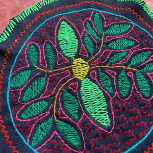 Puede incluir: Un tapiz de pared circular bordado con un diseño floral. El diseño presenta un círculo amarillo central rodeado de hojas verdes, rosas y naranjas. El fondo es negro.