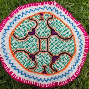 Puede incluir: Un posavasos redondo de tela blanca con un diseño bordado colorido. El diseño presenta un motivo central de cuatro formas conectadas, rodeado por un borde verde y naranja. El posavasos está bordeado con costuras rosas y azules.