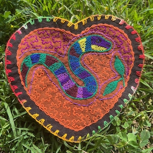May include: Heart-shaped, embroidered textile featuring a colorful snake design. The snake is depicted in shades of blue, green, purple, and yellow, set against a brown and orange background. The edges are trimmed with red, yellow, and black stitching.