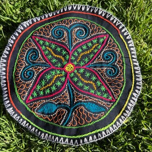 Op de afbeelding: Cirkelvormig zwart geborduurd textiel met een bloemmotief. Het ontwerp heeft een centrale bloem met roze, groene en gele accenten, omgeven door turquoise, oranje en witte borduursels. De rand is afgewerkt met witte en groene stiksels.