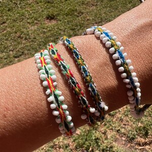 May include: Four woven bracelets featuring intricate patterns in red, green, blue, and yellow thread. Two of the bracelets are embellished with small, round white beads along the edges, while the other two display a braided, multi-coloured textile design.
