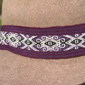 May include: A tan hat with a decorative band. The band is a deep purple color with a white and black geometric pattern. The hat's brim is visible, and the band is wrapped around the base of the crown.
