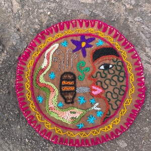 May include: A circular embroidered patch with a colourful design. The centre features a stylised face with a green eye, a pink mouth, and a brown head. The face is surrounded by a green snake, a brown pot, and a purple flower.