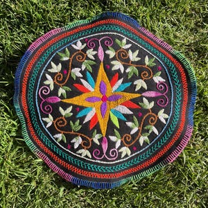 Op de afbeelding: Rond, zwart geborduurd textiel met een kleurrijk sterrenpatroon in het midden. Het sterrenpatroon is omgeven door groene bladeren en ranken. De rand heeft een regenboog van kleuren, waaronder rood, groen en blauw.