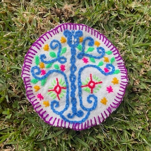Op de afbeelding: Een ronde, witte stoffen patch met een kleurrijk geborduurd ontwerp van een boom met blauwe, groene, roze en gele draad. De patch is omgeven door een paarse rand met een gele en witte afwerking.