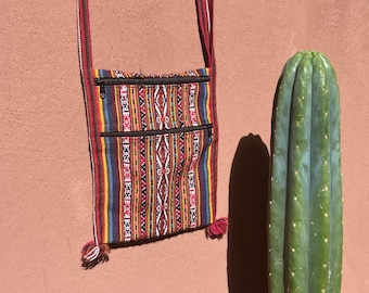 May include: A colorful woven crossbody bag with a geometric pattern in red, blue, yellow, and white. The bag has two zippered pockets and a red strap. A tall green cactus stands to the right against a brown wall.