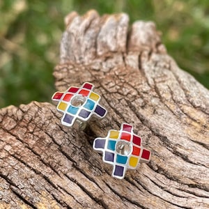 May include: A pair of silver-toned stud earrings with a colorful mosaic design. Each earring features a square pattern with red, yellow, blue, and purple squares. The earrings are set against a textured wooden surface.