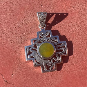 May include: Silver pendant with a cross design, featuring a central round, light green stone. The pendant has intricate details, including animal and bird motifs, and is suspended from a decorative bail. The pendant is displayed on a red surface.