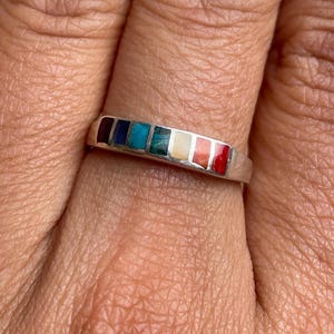 Può includere: Un anello a fascia d'argento con una fila di pietre multicolori incastonate. Le pietre sono disposte in fila e sono di colore rosso, arancione, giallo, verde, blu e viola.