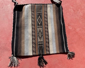 May include: A woven tapestry bag with brown, black, and white stripes and geometric patterns. The bag has fringe tassels on the bottom.