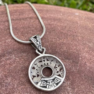 May include: Silver-toned pendant necklace with a circular pendant featuring an open center and intricate cut-out designs. The pendant is attached to a thin, silver chain. The necklace is displayed on a reddish-brown surface.