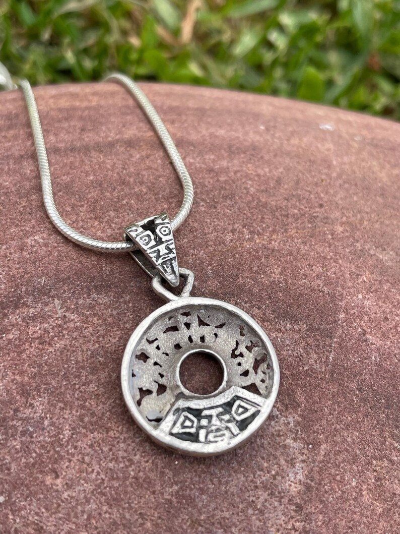 May include: Silver-toned pendant necklace with a circular pendant featuring an open center and intricate cut-out designs. The pendant is attached to a thin, silver chain. The necklace is displayed on a reddish-brown surface.