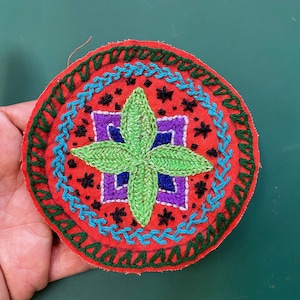 May include: A round, red fabric patch with a green, four-pointed star in the center. The star is surrounded by a purple and blue square design. The patch is edged with a green, blue, and red stitched border.