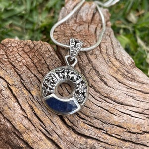 May include: A silver pendant necklace with a circular design and a dark blue stone inlay. The pendant is on a silver chain and rests on a piece of weathered wood. The pendant has intricate details.