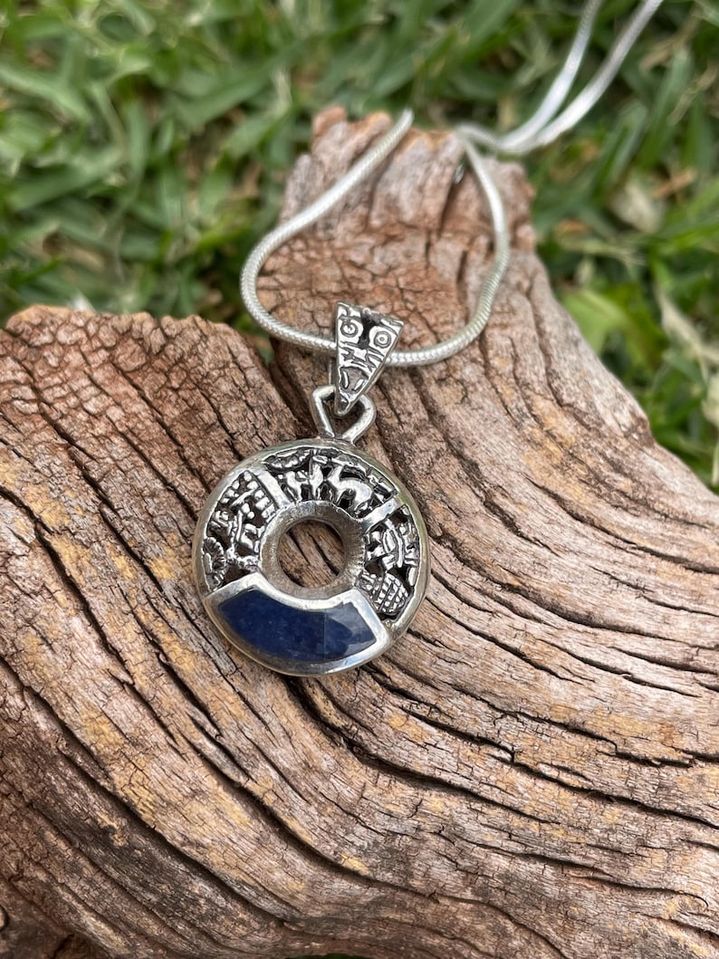 May include: A silver pendant necklace with a circular design and a dark blue stone inlay. The pendant is on a silver chain and rests on a piece of weathered wood. The pendant has intricate details.