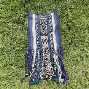 May include: A woven bag with a blue, white, and black geometric pattern. The bag has a long, thin strap and a fringe at the bottom. The bag is lying on green grass.