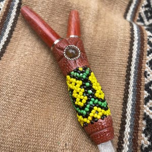 May include: A handcrafted wooden pipe with a Y-shaped design. The pipe features a carved brown body adorned with yellow, green, and black beaded patterns. A clear crystal is set at the base, and a gemstone is set in the center.