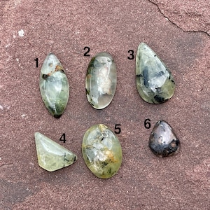 May include: Six green and black gemstone cabochons of various shapes and sizes. The cabochons are arranged in a row on a brown, textured surface.