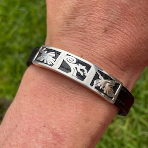 May include: A black leather bracelet with silver metal accents featuring four different designs. The designs are stylized and abstract.