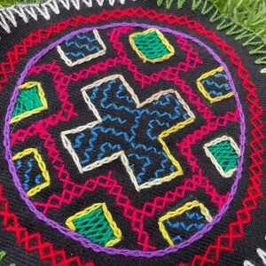 Puede incluir: Una pieza circular de tela negra con bordados coloridos. El bordado presenta un diseño en forma de cruz con patrones geométricos en tonos de azul, verde, amarillo, rosa y blanco.
