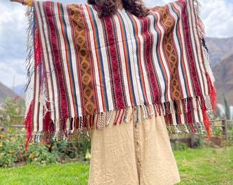 Könnte beinhalten: Ein farbenfrohes gewebtes Poncho mit vertikalen Streifen in Rot, Weiß und Blau und einem Fransenbesatz. Der Poncho wird über einem beigefarbenen Rock getragen. Die Person trägt einen braunen Hut und Stiefel. Der Hintergrund umfasst grünes Gras und einen Berg.