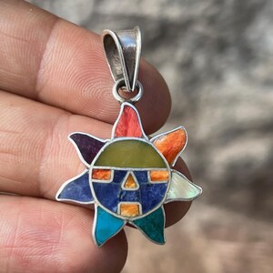 Puede incluir: Un colgante de plata con forma de sol con detalles incrustados de colores. El diseño del sol presenta una cara con formas geométricas en tonos azules, naranjas, verdes y amarillos. El colgante está sujeto por una mano.