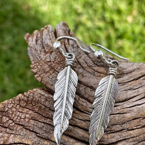 May include: A pair of silver-colored feather earrings. Each earring features a detailed feather design suspended from a curved ear wire. The earrings are displayed against a natural wood background with a blurred green backdrop.
