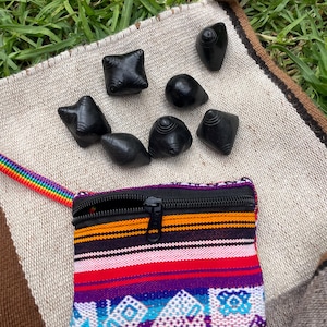 Può includere: Una collezione di sette piccoli oggetti in pietra nera scolpita di varie forme, appoggiati su un tessuto beige. È presente anche una piccola pochette rettangolare con cerniera con un colorato motivo a righe e un cinturino arcobaleno.