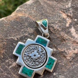 May include: A silver pendant with a cross-shaped design, featuring a central circular emblem with intricate patterns. The pendant is adorned with rectangular turquoise inlays and a teardrop-shaped turquoise stone at the top. The pendant is displayed on a rough, gray stone surface.