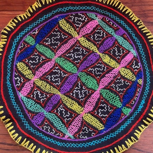Puede incluir: Un posavasos redondo de tela negra con un diseño bordado de colores. El diseño presenta un patrón repetitivo de óvalos amarillos, rosas, verdes, azules y morados.