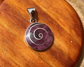 May include: A circular silver pendant featuring a deep purple stone inlay arranged in a spiral pattern. The pendant includes a silver bail at the top with a small, dark, teardrop-shaped stone accent, creating a cohesive and detailed jewelry piece.