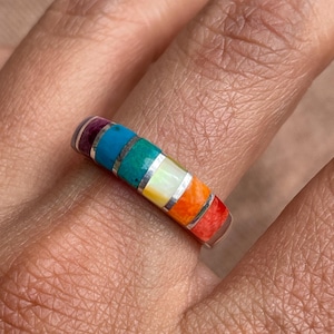 Op de afbeelding: Een zilveren ring met een regenboog van ingelegde stenen. De stenen zijn in een band rond de ring gerangschikt en omvatten paars, turquoise, groen, wit, geel, oranje en rood.