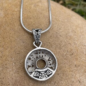 May include: A silver pendant necklace featuring a circular pendant with an intricate, openwork design. The pendant has a central hole and is suspended from a delicate silver chain. The pendant's design includes geometric and stylized patterns.
