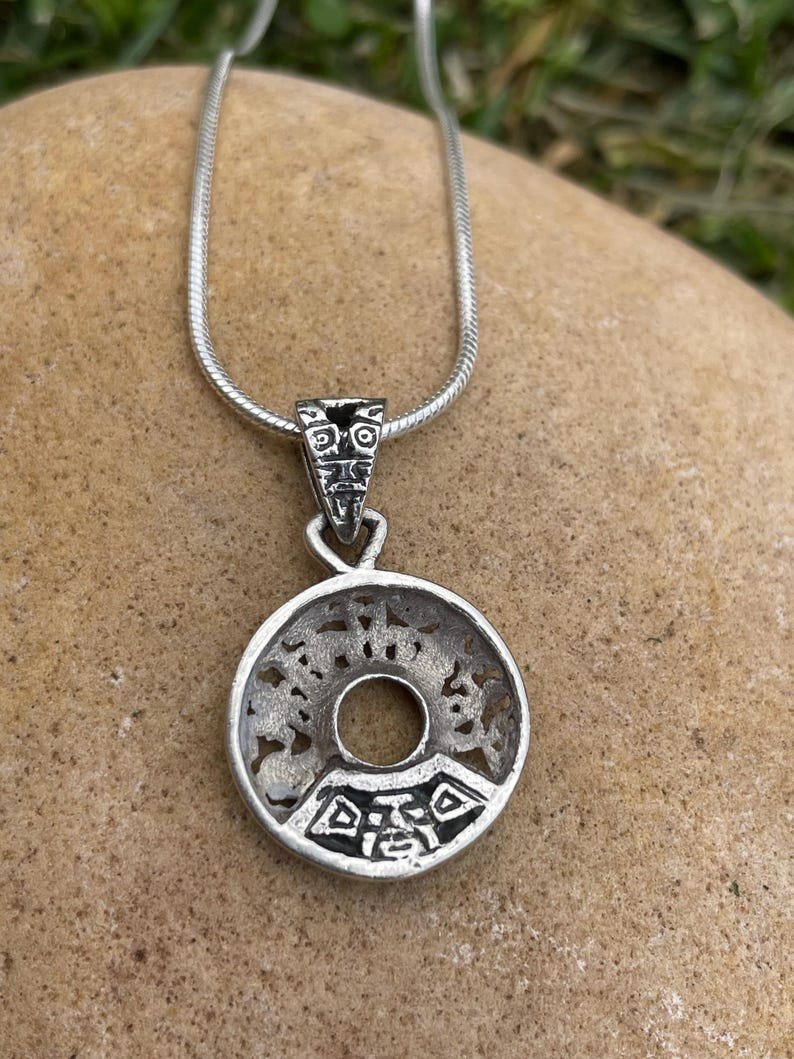 May include: A silver pendant necklace featuring a circular pendant with an intricate, openwork design. The pendant has a central hole and is suspended from a delicate silver chain. The pendant's design includes geometric and stylized patterns.