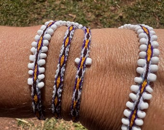 May include: A multi-wrap bracelet featuring a woven pattern in yellow, purple, and white thread. The design is accented with small, opaque white beads lining the edges of the woven band, creating a textured appearance around the wrist.