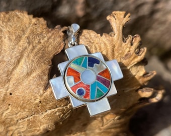 May include: A silver pendant with a colorful, geometric design. The pendant features a central circle with a mosaic of red, blue, green, and purple shapes, surrounded by a white cross-shaped frame. The pendant is suspended from a silver chain.