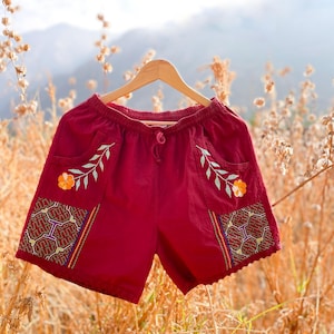 May include: Burgundy cotton shorts with embroidered floral and geometric designs. The shorts have a drawstring waist and are hanging on a wooden hanger.