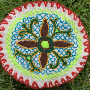 Op de afbeelding: Een ronde geborduurde patch met een witte achtergrond en een kleurrijk bloemenontwerp. Het ontwerp heeft een bruine vierpuntige ster met groene bladeren en blauwe en gele swirls.
