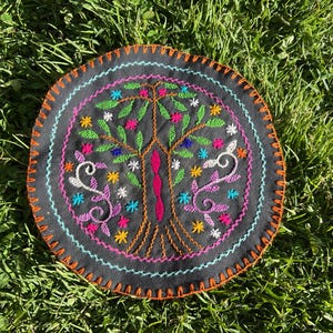 May include: Round, black embroidered textile with a colorful tree design. The tree features green leaves, pink and blue flowers, and an orange trunk. The border is orange with a blue and pink stitch.