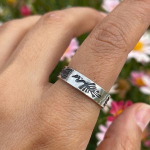 Peut inclure: Une bague en argent avec un motif d'oiseau détaillé. La bague est portée au doigt, mettant en valeur le motif gravé complexe. L'oiseau est représenté avec les ailes déployées. La conception de la bague est un bijou saisissant.