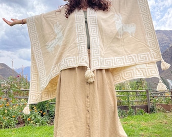 May include: A beige poncho with white geometric border and llama design. The poncho has tassels and is worn with a matching skirt and brown boots. The outfit is set against a backdrop of green grass and a cloudy sky.