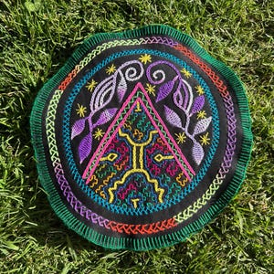 Op de afbeelding: Ronde, zwart geborduurde patch met een kleurrijk geometrisch ontwerp. De patch heeft een centrale driehoek, bloemaccenten en een rand van verschillende gekleurde steken. De achtergrond is groen gras.