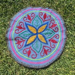 Puede incluir: Textil bordado azul de forma redonda con un diseño floral. El centro presenta una flor amarilla y azul, rodeada de hojas verdes y detalles rojos rizados. El borde está adornado con costuras marrones, detalles rosas y verdes.