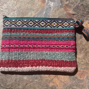 May include: A rectangular zippered pouch featuring a woven textile pattern with horizontal stripes in shades of magenta, burgundy, sage green, and cream. The top border displays a black band with a white, yellow, and teal geometric diamond motif.
