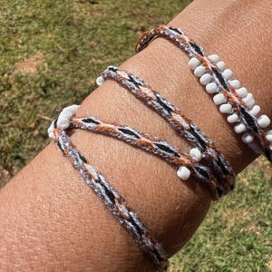 May include: A multi-wrap bracelet constructed from braided thread in shades of black, white, and orange. The design features small white cylindrical beads woven along the length of the cord, creating a textured appearance across the wrist.