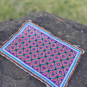 May include: A small, rectangular rug with a geometric pattern in shades of red, orange, green, blue, and purple. The rug has a white background and a fringed edge.
