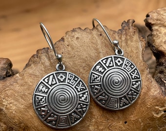 May include: A pair of silver earrings with a circular design featuring a spiral pattern and intricate symbols around the edge.